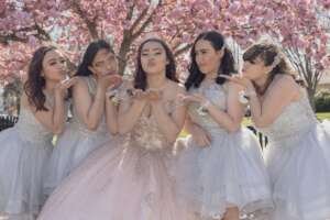 Five young women celebrate a quinceañera under blooming trees in elegant dresses, exuding joy and friendship.